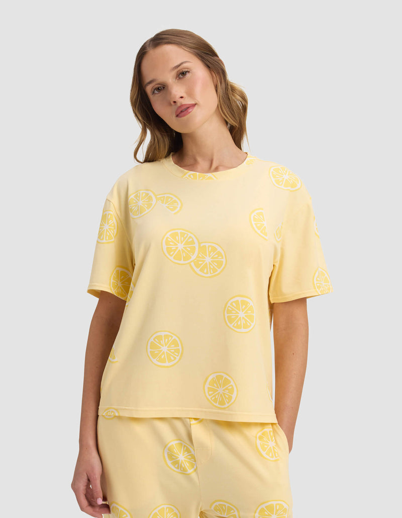 A woman wears the Cozy Earth Women's First Layer Pajama Short Sleeve Tee, paired with matching lemon slice patterned pants, standing against a plain light gray background.