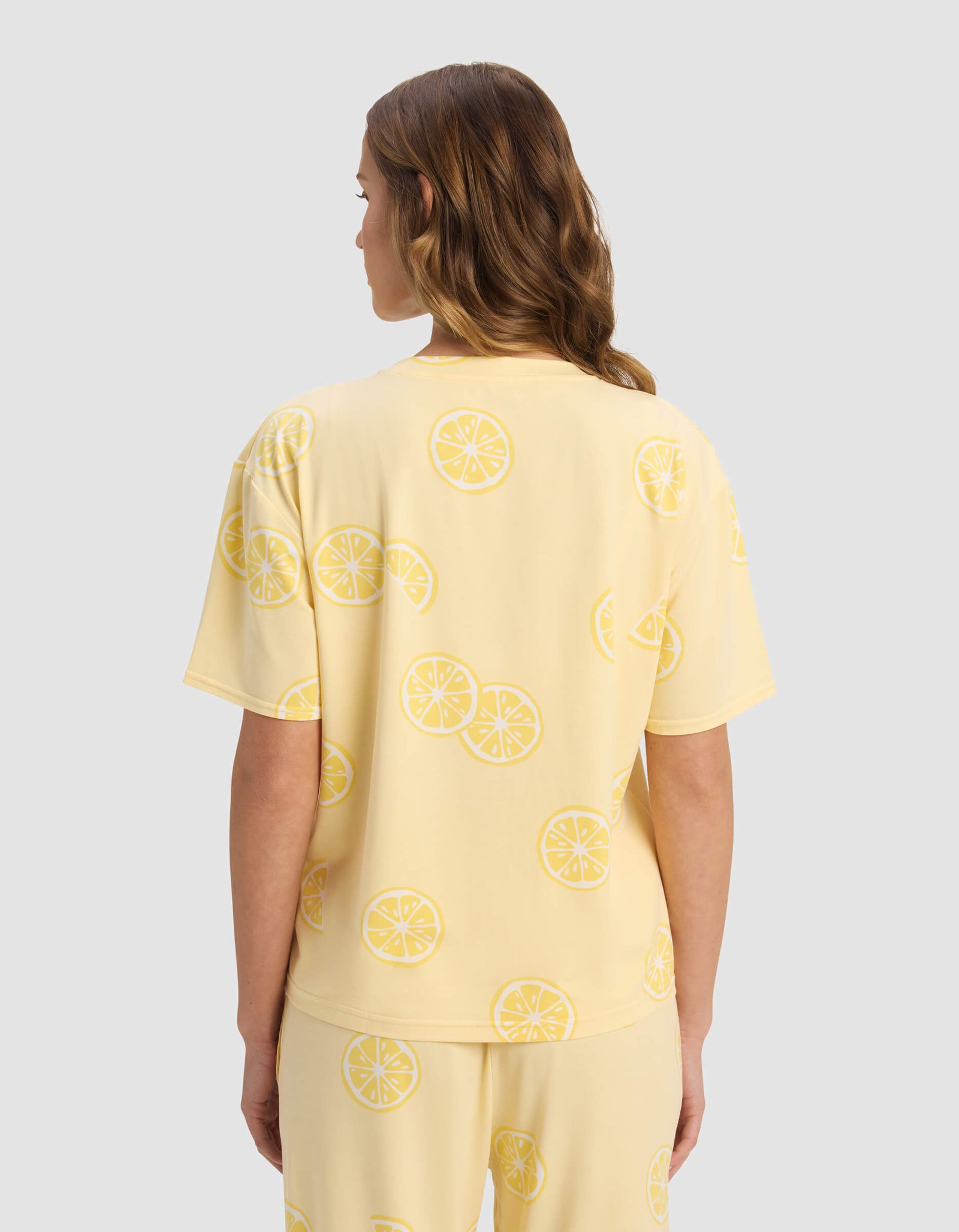 A woman with wavy, light brown hair is seen from the back wearing Cozy Earth's Women's First Layer Pajama Short Sleeve Tee in a lemon slice print set, standing against a plain light gray background.