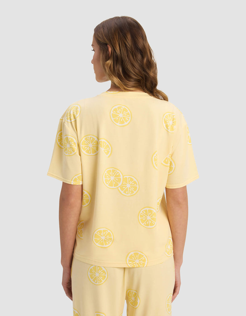 A woman with wavy, light brown hair is seen from the back wearing Cozy Earth's Women's First Layer Pajama Short Sleeve Tee in a lemon slice print set, standing against a plain light gray background.