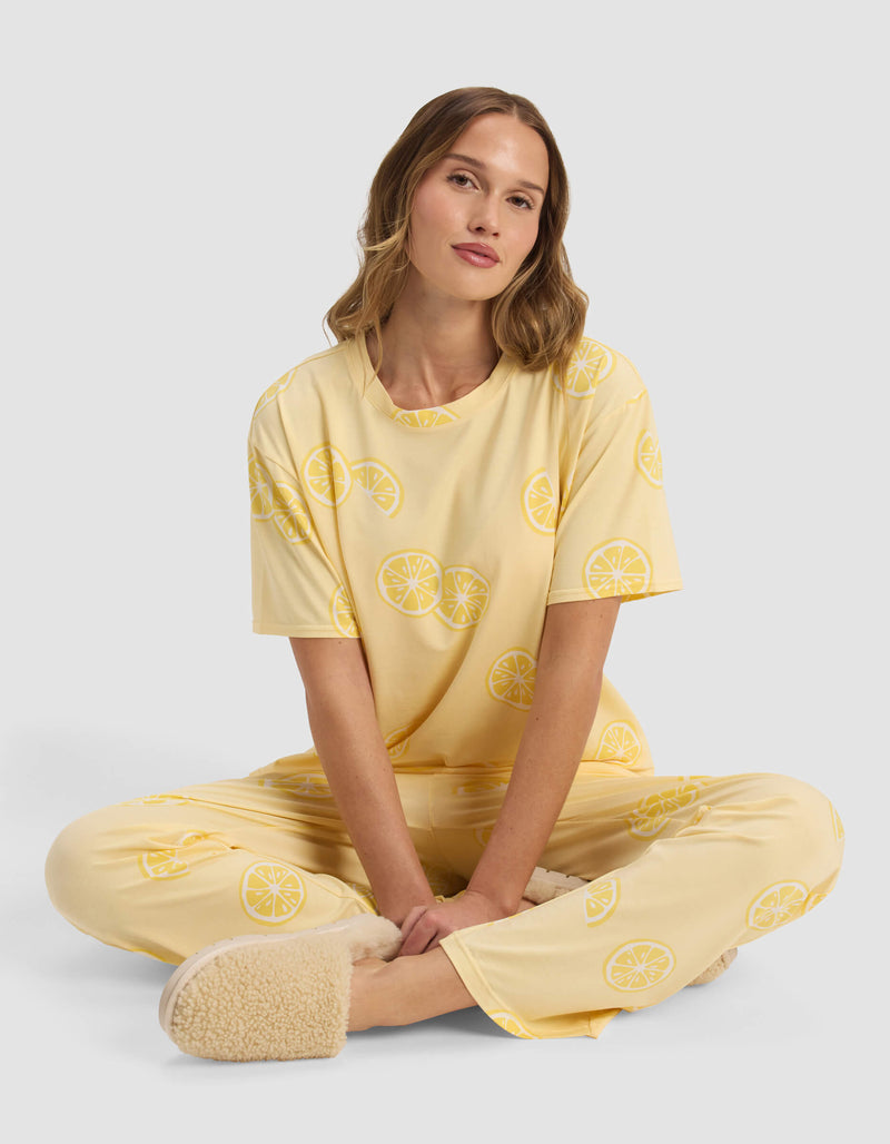 A woman in Cozy Earth’s Women’s First Layer Pajama Short Sleeve Tee sits cross-legged on the floor against a light background, wearing lemon-patterned yellow pajamas and fuzzy beige slippers.