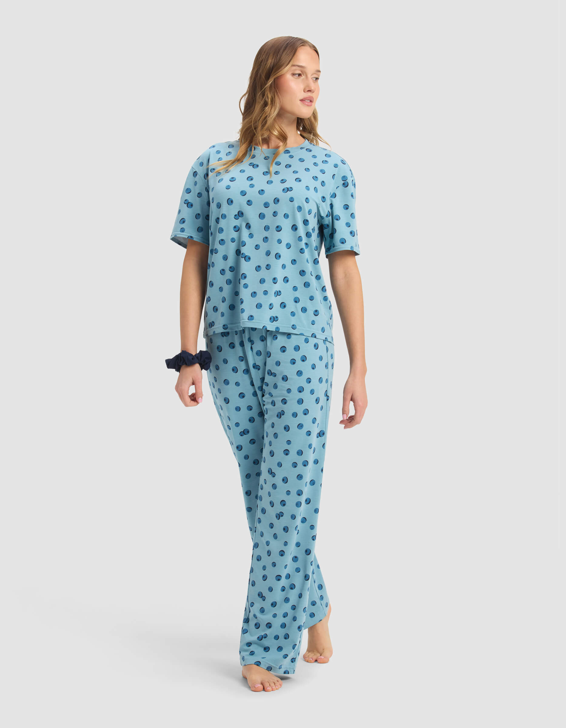 A woman in Cozy Earth's Women's First Layer Pajama Short Sleeve Tee & Pant Set, light blue with dark blue dots, walks barefoot against a plain backdrop. She wears a dark scrunchie on her wrist and has loose hair.