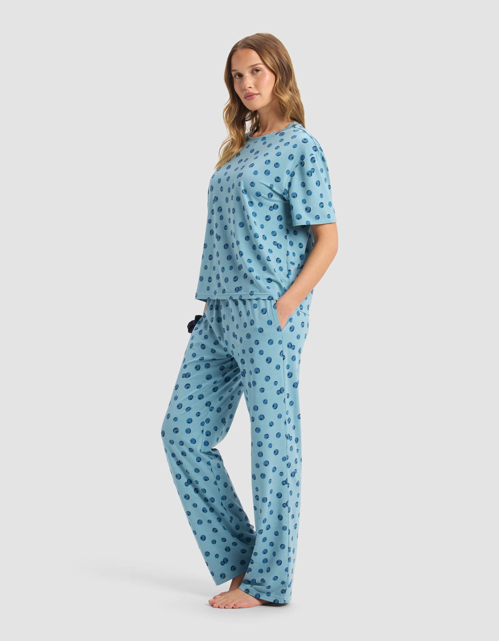 A woman stands barefoot, facing sideways and looking at the camera, wearing Cozy Earth’s Women’s First Layer Pajama Short Sleeve Tee & Pant Set in light blue with a dark blue polka dot pattern.