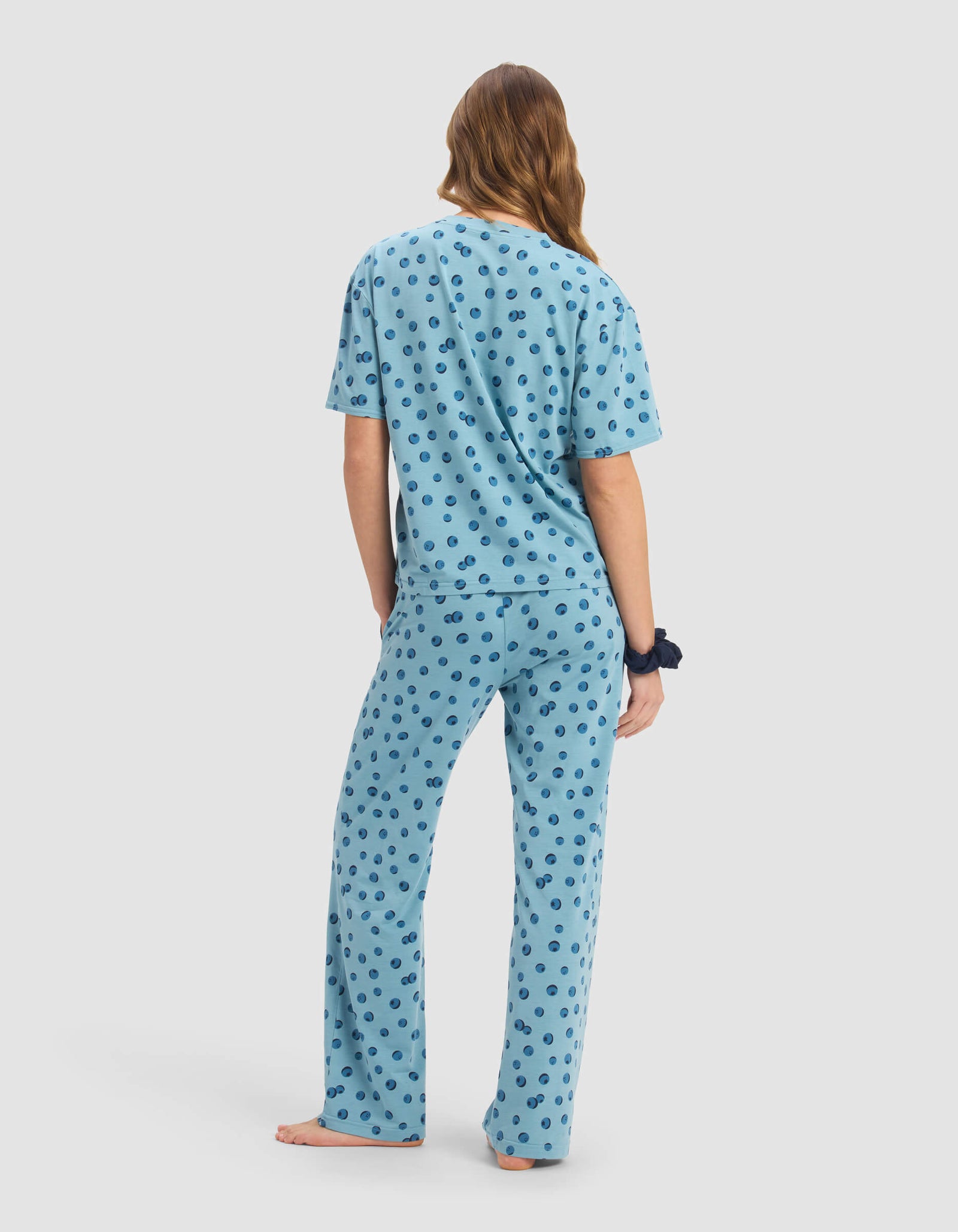 A person with long hair stands barefoot and facing away, wearing the Cozy Earth Women's First Layer Pajama Short Sleeve Tee & Pant Set in light blue with dark smiley face patterns, and a navy scrunchie on their left wrist.