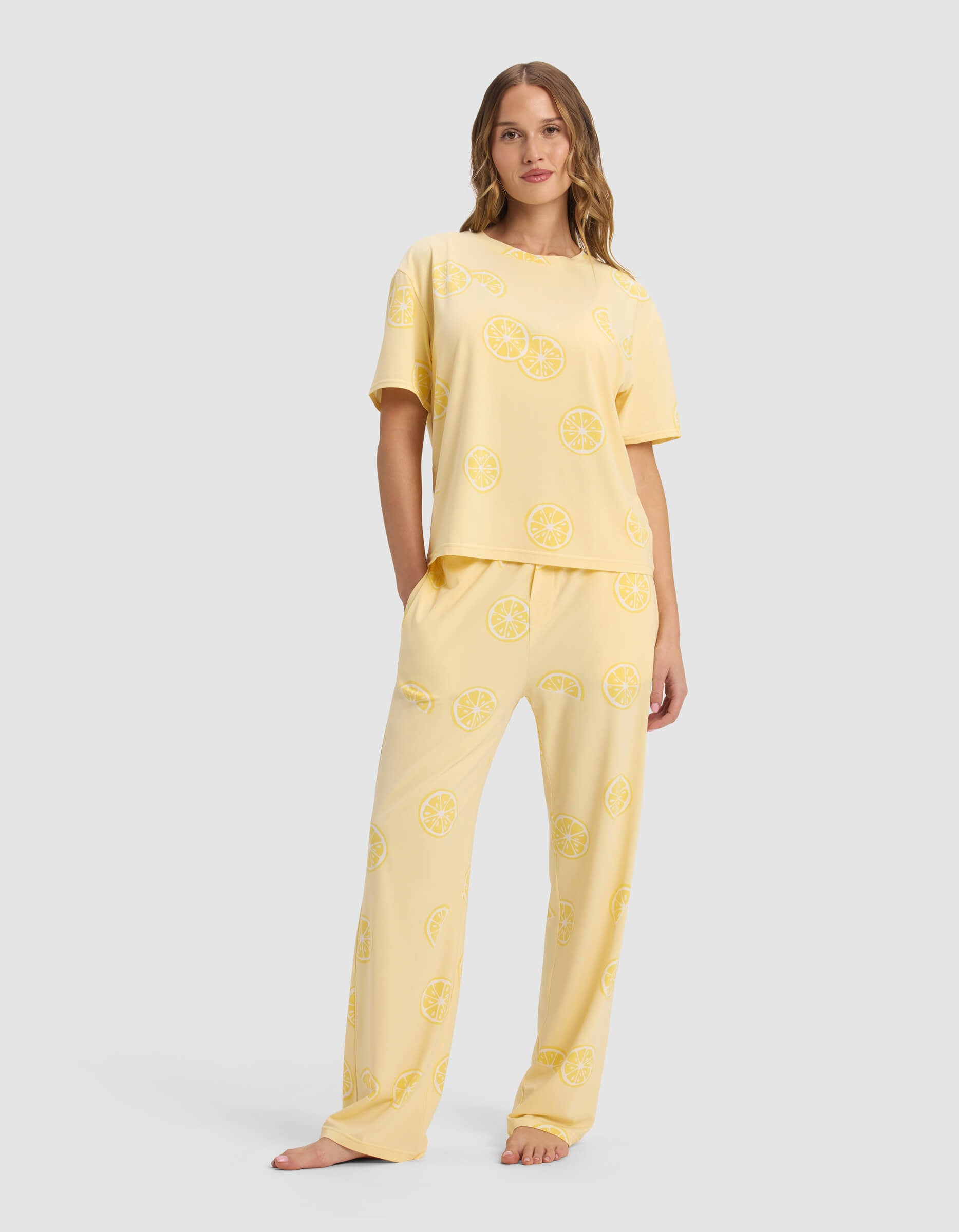 A woman with long hair and a neutral expression stands barefoot wearing Cozy Earth’s Women's First Layer Pajama Short Sleeve Tee & Pant Set, featuring a relaxed fit and a sliced lemon print on light yellow fabric.