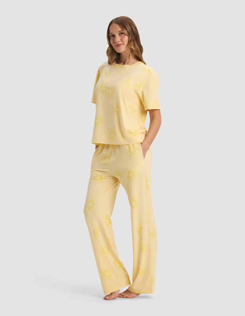 A woman stands barefoot, wearing the Cozy Earth Women's First Layer Pajama Short Sleeve Tee & Pant Set in light yellow, featuring a loose fit and an all-over yellow lemon slice print.