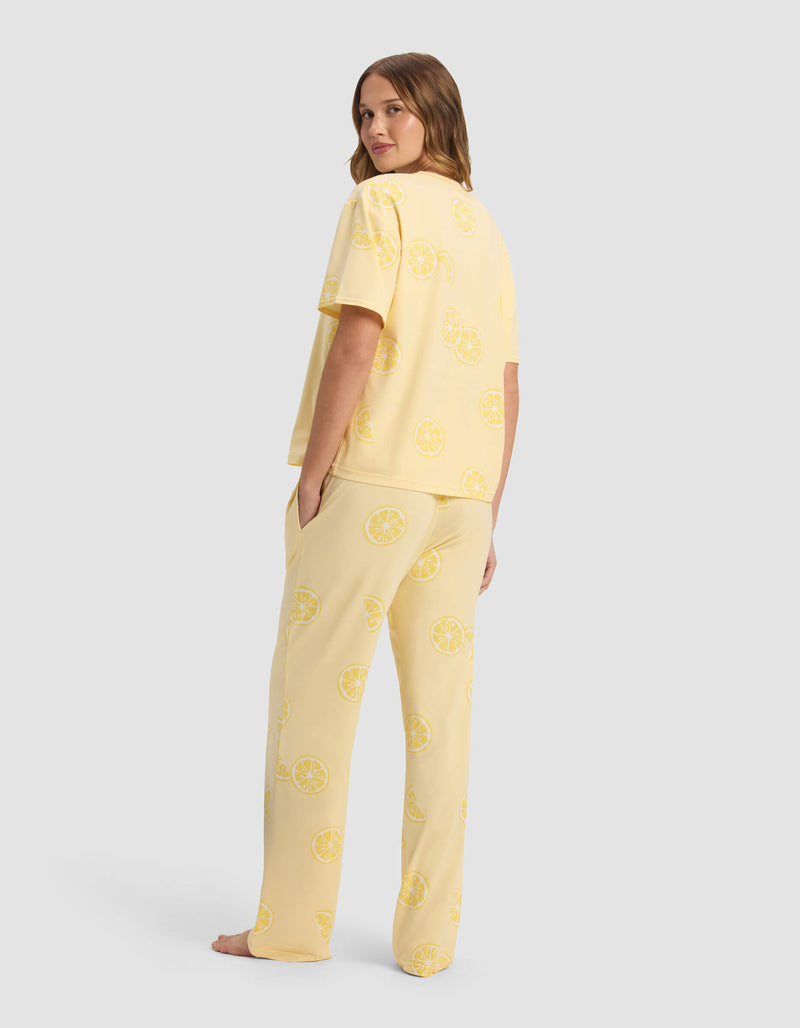 A woman stands barefoot, facing backward and looking over her shoulder, wearing the Cozy Earth Women's First Layer Pajama Short Sleeve Tee & Pant Set with a yellow lemon slice pattern against a plain light gray background.