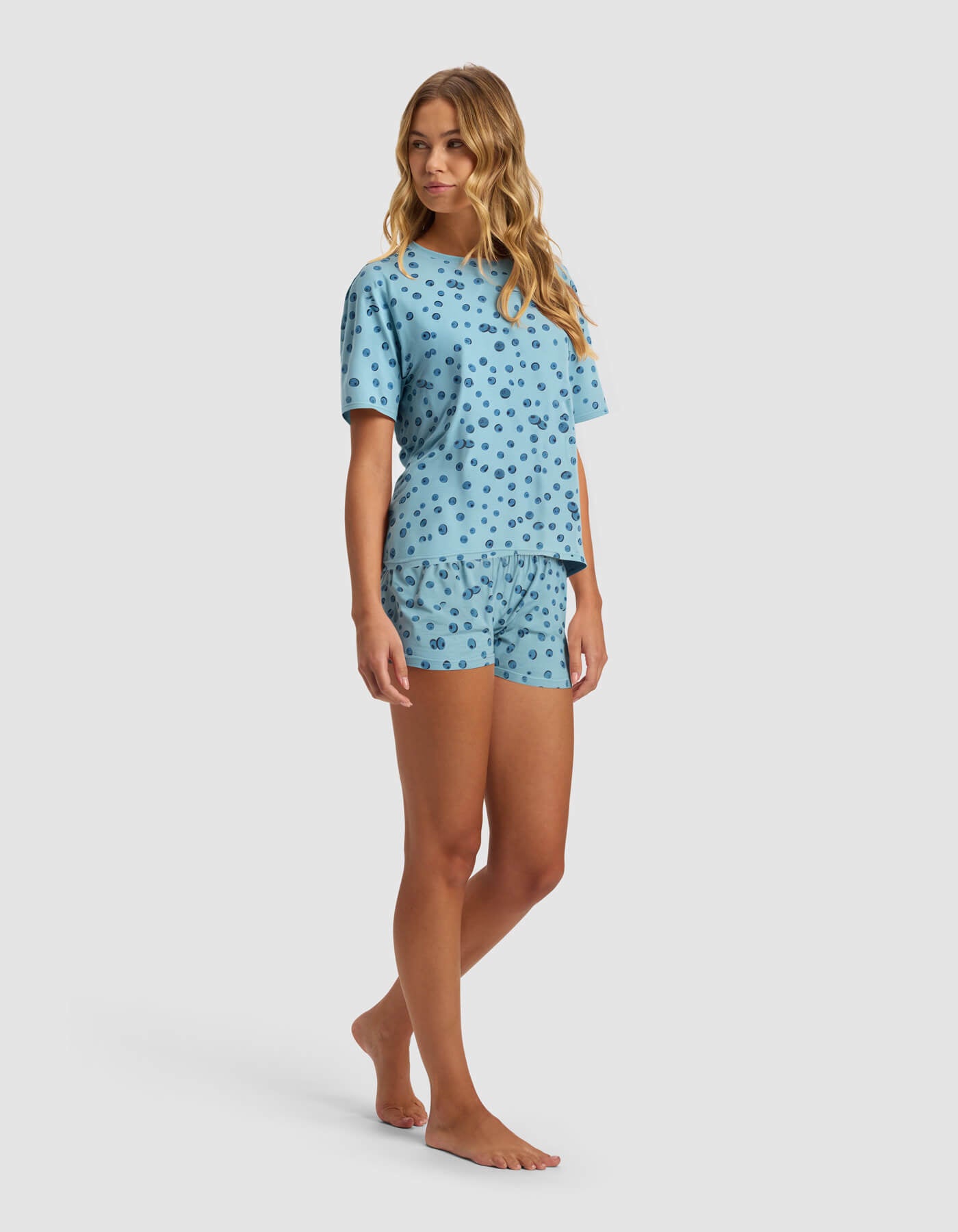 A woman stands barefoot against a white background wearing the Cozy Earth Women's First Layer Pajama Short Sleeve Tee & Boxer Short Set in light blue with dark blue polka dots.