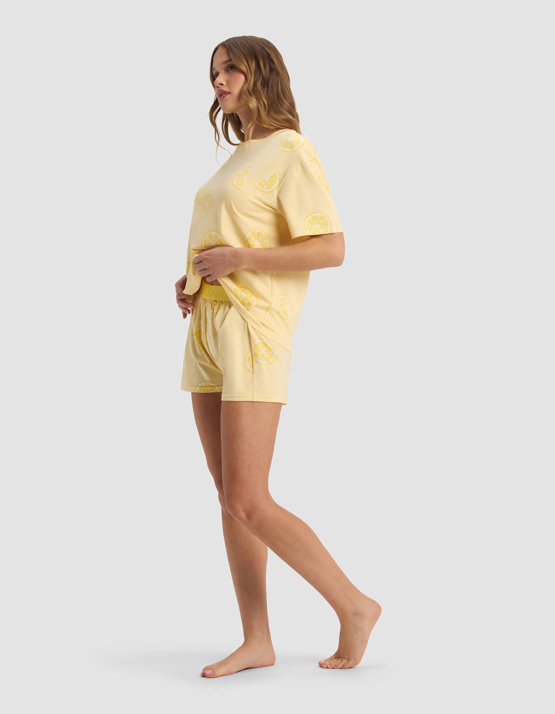 A woman stands barefoot in a Cozy Earth Women's First Layer Pajama Short Sleeve Tee & Boxer Short Set, featuring a lemon slice print and light yellow color, facing sideways against a plain light background.