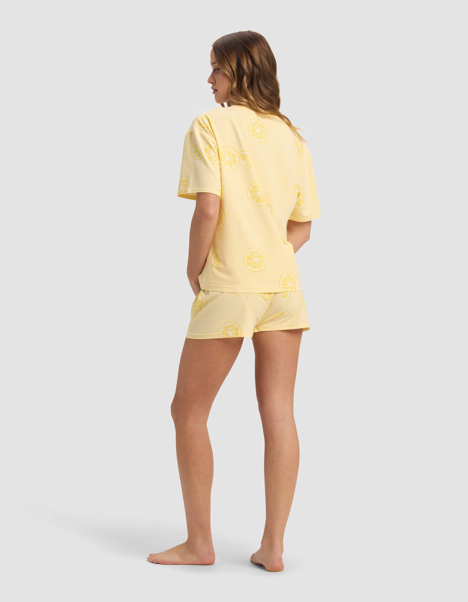 A woman stands barefoot, facing away, wearing Cozy Earth's Women's First Layer Pajama Short Sleeve Tee & Boxer Short Set in yellow citrus print. Her hands are in her shorts' pockets, hair loose, against a plain light gray background.
