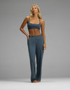 A woman stands barefoot against a plain gray background, wearing the Cozy Earth Women’s First Layer Pajama Soft Bra & Pajama Pant Set in teal. She has long blonde hair and is posing with one hand in her pocket and the other resting at her side.|Color:Dawn