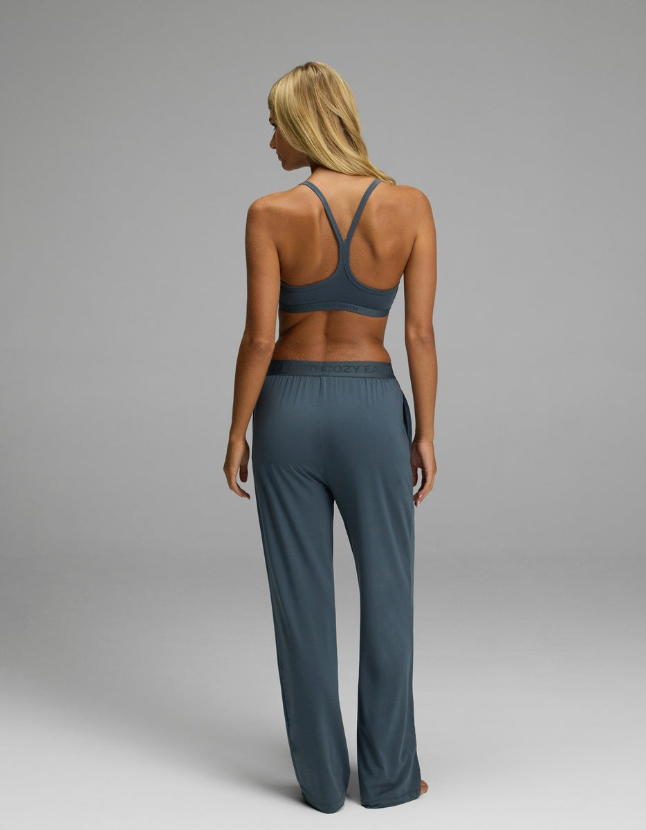 A woman with blonde hair, seen from behind, models the Cozy Earth Women’s First Layer Pajama Soft Bra & Pajama Pant Set in blue. She stands barefoot against a plain gray background.
