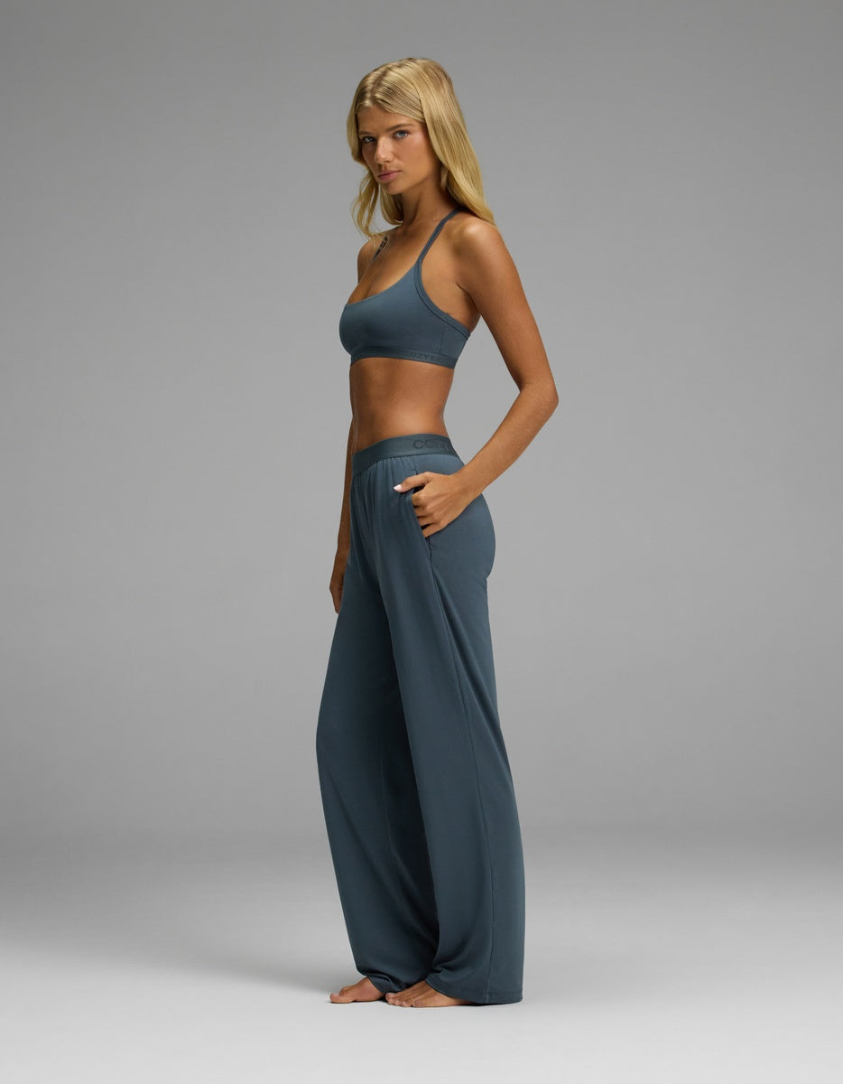 A woman stands barefoot in a studio, modeling the Cozy Earth Women’s First Layer Pajama Soft Bra & Pajama Pant Set in matching blue. She faces sideways with one hand in her pocket, her blonde hair down, set against a plain grey background.