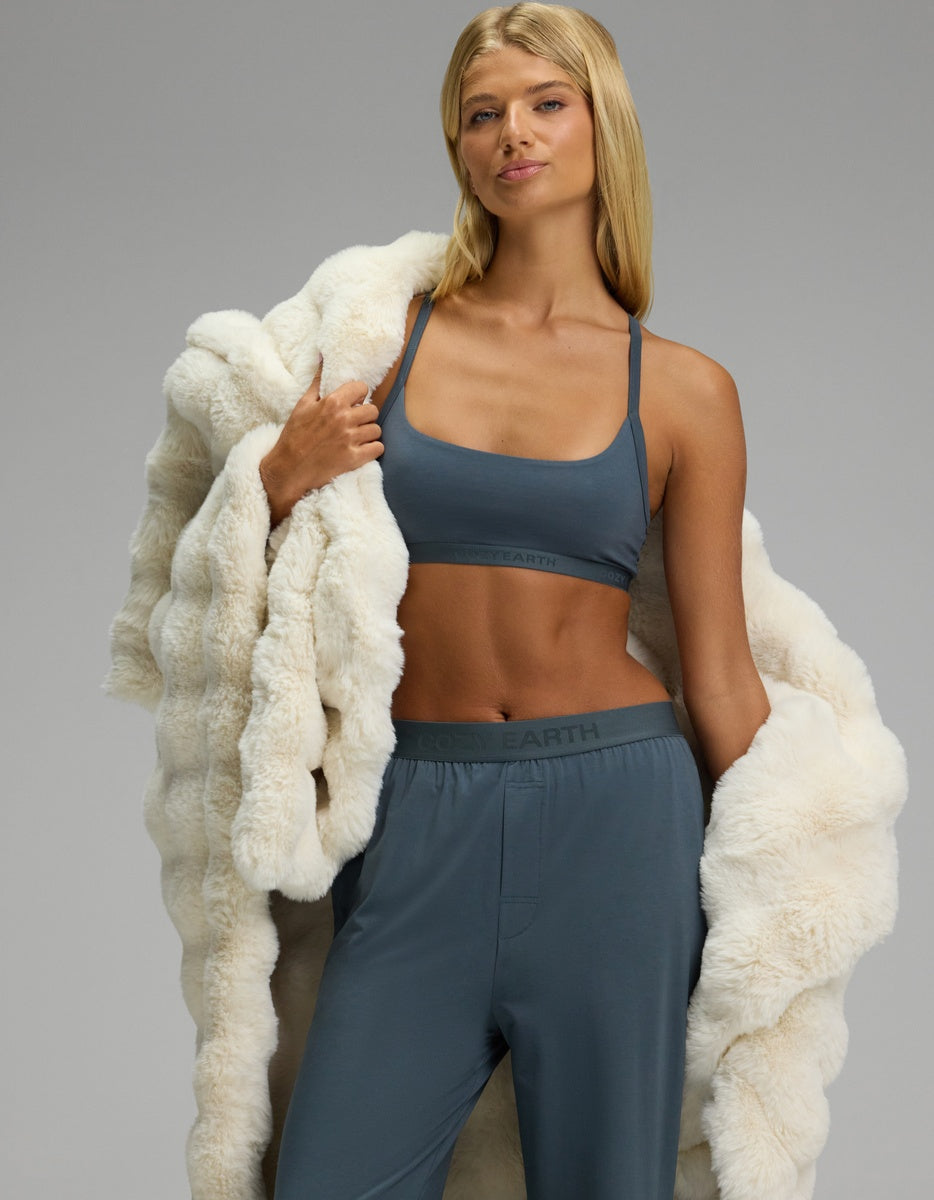 A woman models the Cozy Earth Women’s First Layer Pajama Soft Bra & Pajama Pant Set, confidently posing with a plush white fur coat draped over her shoulders against a plain gray background.