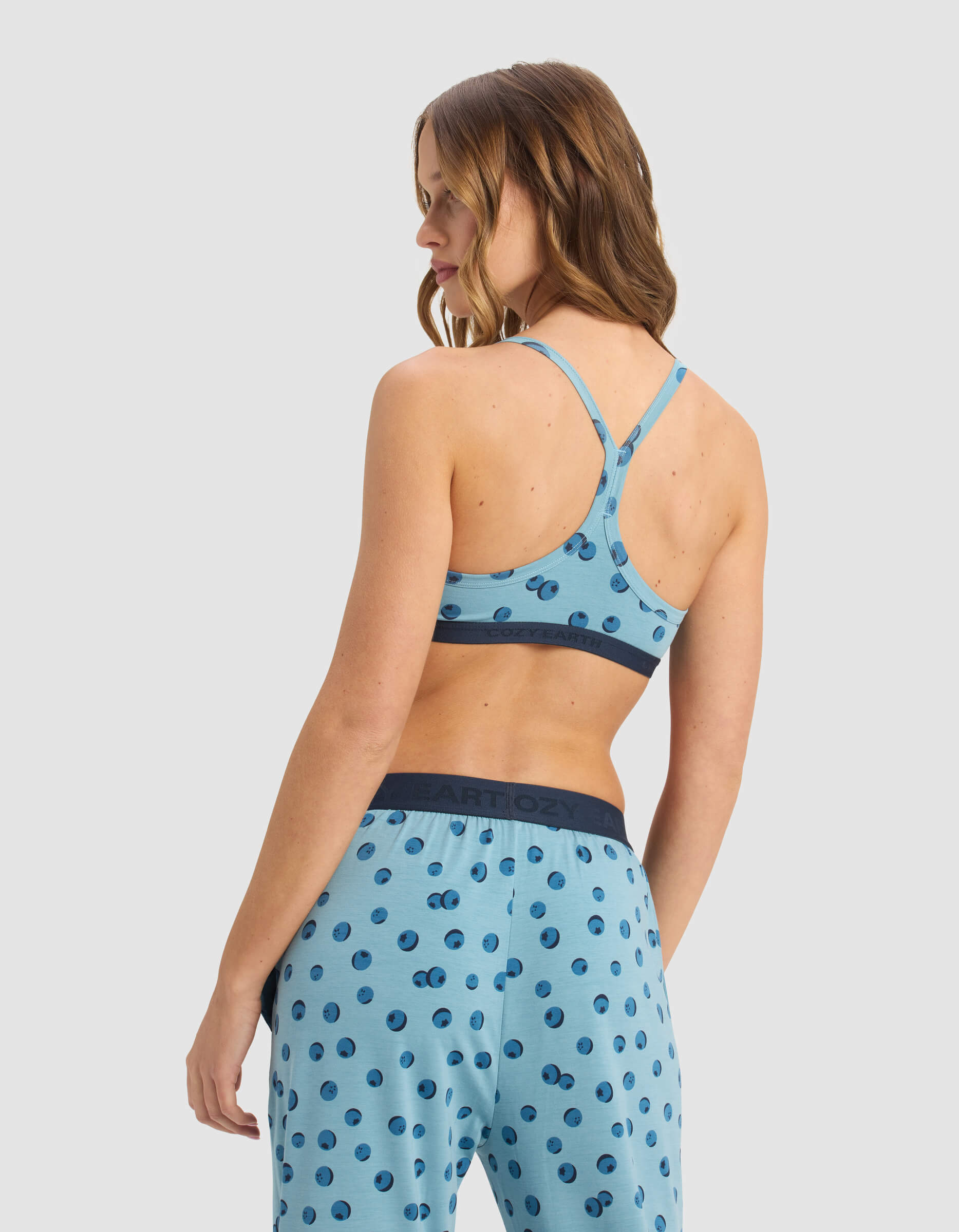 A woman faces away wearing Cozy Earth’s Women’s First Layer Pajama Soft Bra in light blue and matching polka dot pants, featuring a racerback and branded waistband against a plain, light background.