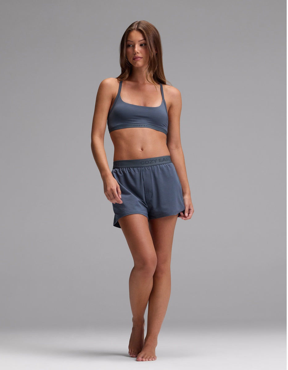 A woman stands barefoot against a plain background, wearing the Cozy Earth Women’s First Layer Pajama Soft Bra & Boxer Short Set in Dawn. She has long brown hair and a neutral expression.