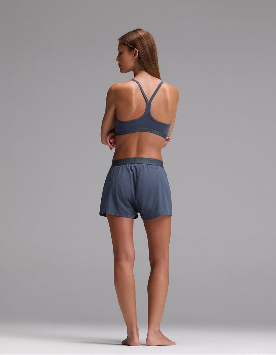 A woman stands barefoot on a gray floor, facing away, wearing the Cozy Earth Women’s First Layer Pajama Soft Bra & Boxer Short Set in blue. The background is plain and gray.