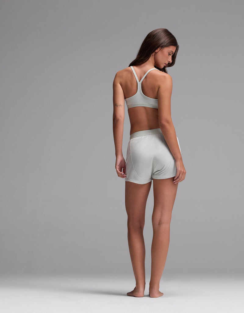 A woman stands barefoot on a gray background, facing away while wearing the Cozy Earth Women’s First Layer Pajama Soft Bra & Boxer Short Set. She looks over her shoulder with her arms relaxed by her sides.