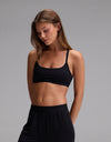 A woman with long brown hair wears the Cozy Earth Women’s First Layer Soft Bra in Jet Black, standing against a plain gray background and looking down with a neutral expression.|Color:Jet Black