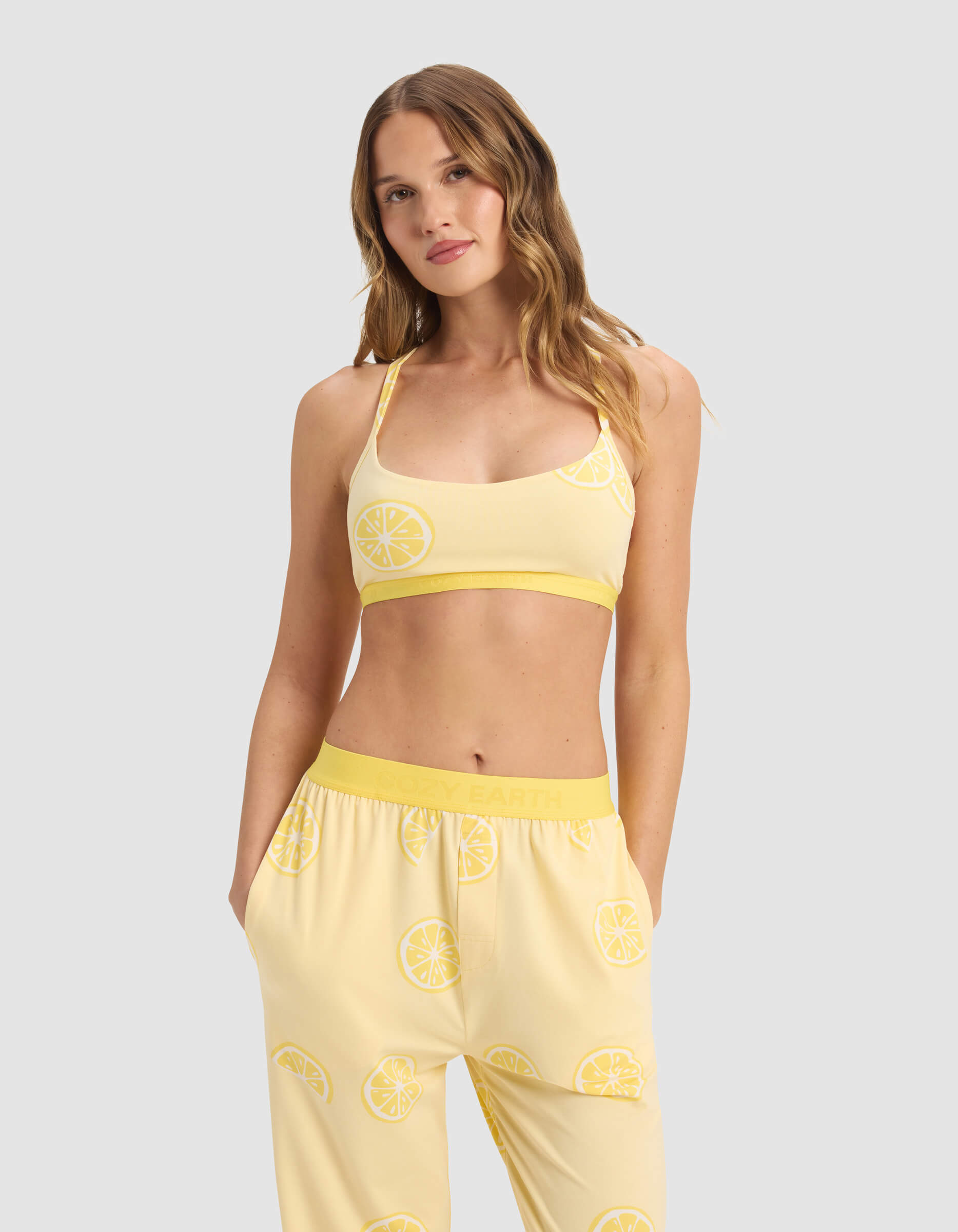 A woman with wavy, light brown hair wears the Cozy Earth Women's First Layer Pajama Soft Bra and matching lemon print pants, standing against a plain light gray background.|Color:Lemons
