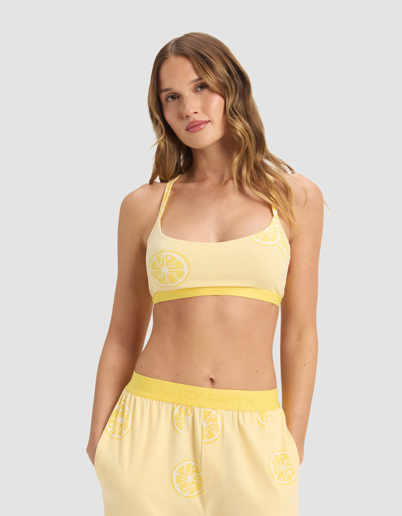A woman with wavy, light brown hair wears the Cozy Earth Women's First Layer Pajama Soft Bra and matching lemon print pants, standing against a plain light gray background.