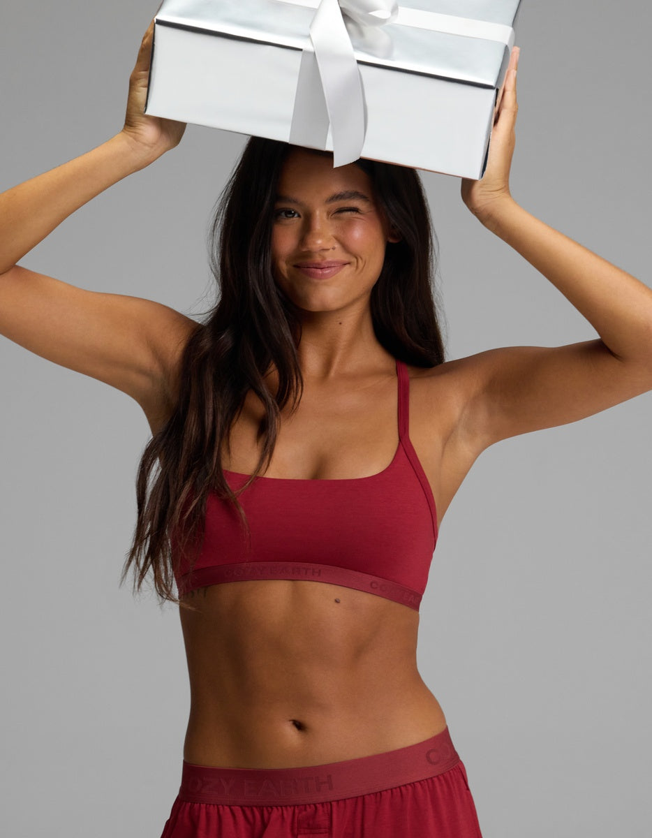 A smiling woman winks while holding a gift box above her head, wearing a burgundy Women’s First Layer Pajama Soft Bra & Pajama Pant Set.