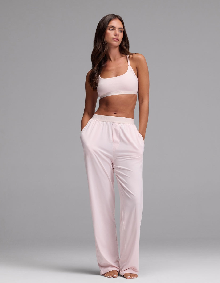 A woman stands with her hands in her pockets against a plain gray background, wearing the Cozy Earth Women’s First Layer Pajama Soft Bra & Pajama Pant Set in Ballet.
