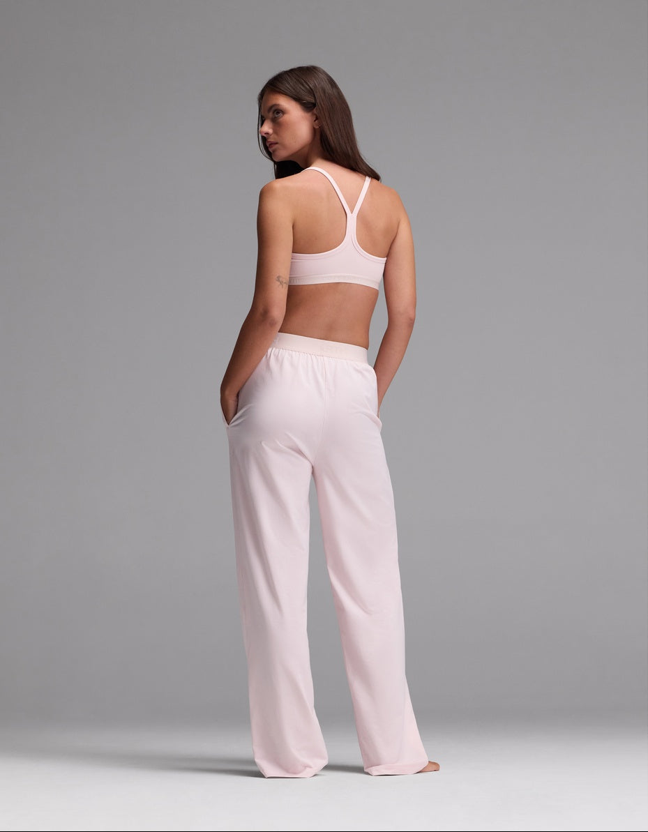 A woman stands with her back to the camera, wearing the Cozy Earth Women’s First Layer Pajama Soft Bra & Pajama Pant Set in light pink. Her hands rest in her pockets as she looks over her shoulder against a plain gray background.