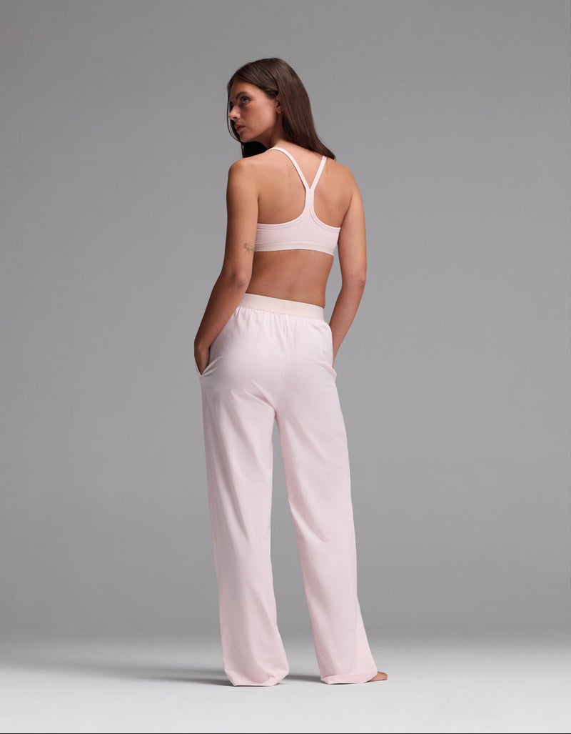 A woman stands with her back to the camera, wearing the Cozy Earth Women’s First Layer Pajama Soft Bra & Pajama Pant Set in light pink. Her hands rest in her pockets as she looks over her shoulder against a plain gray background.