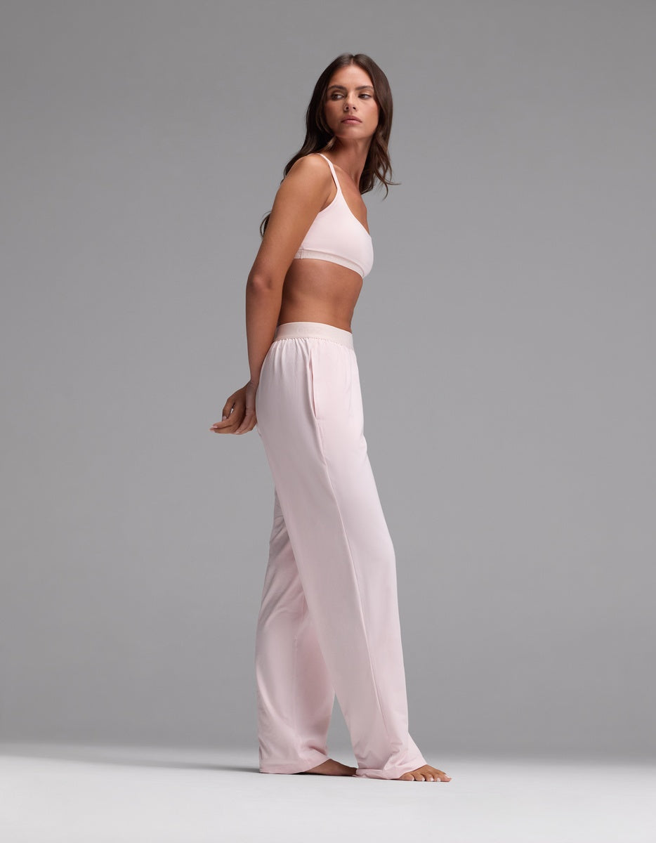 A woman stands barefoot in a studio, wearing the Cozy Earth Women’s First Layer Pajama Soft Bra & Pajama Pant Set in light pink. She looks slightly to the side with her arms behind her back, set against a plain gray background.