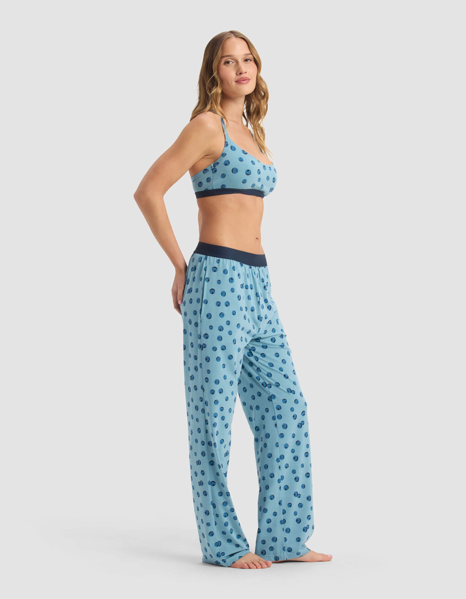 A woman models the Cozy Earth Women's First Layer Pajama Soft Bra & Pant Set in light blue with dark blue polka dots, featuring a matching bralette and loose-fitting pants, posing barefoot against a plain white background.