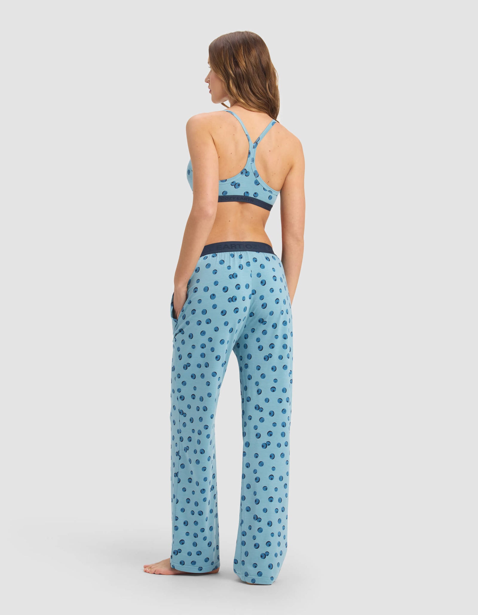 A woman stands barefoot, facing away, wearing Cozy Earth's Women's First Layer Pajama Soft Bra & Pant Set in light blue with a dark blue polka dot pattern. The background is plain and light colored.