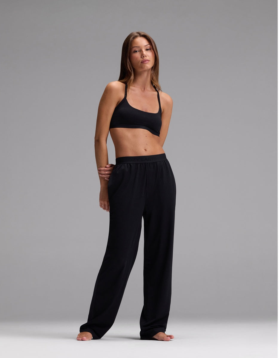 A woman stands barefoot against a gray background, wearing the Cozy Earth Women’s First Layer Pajama Soft Bra & Pajama Pant Set in Jet Black, with one hand at her side and the other bent at her elbow.