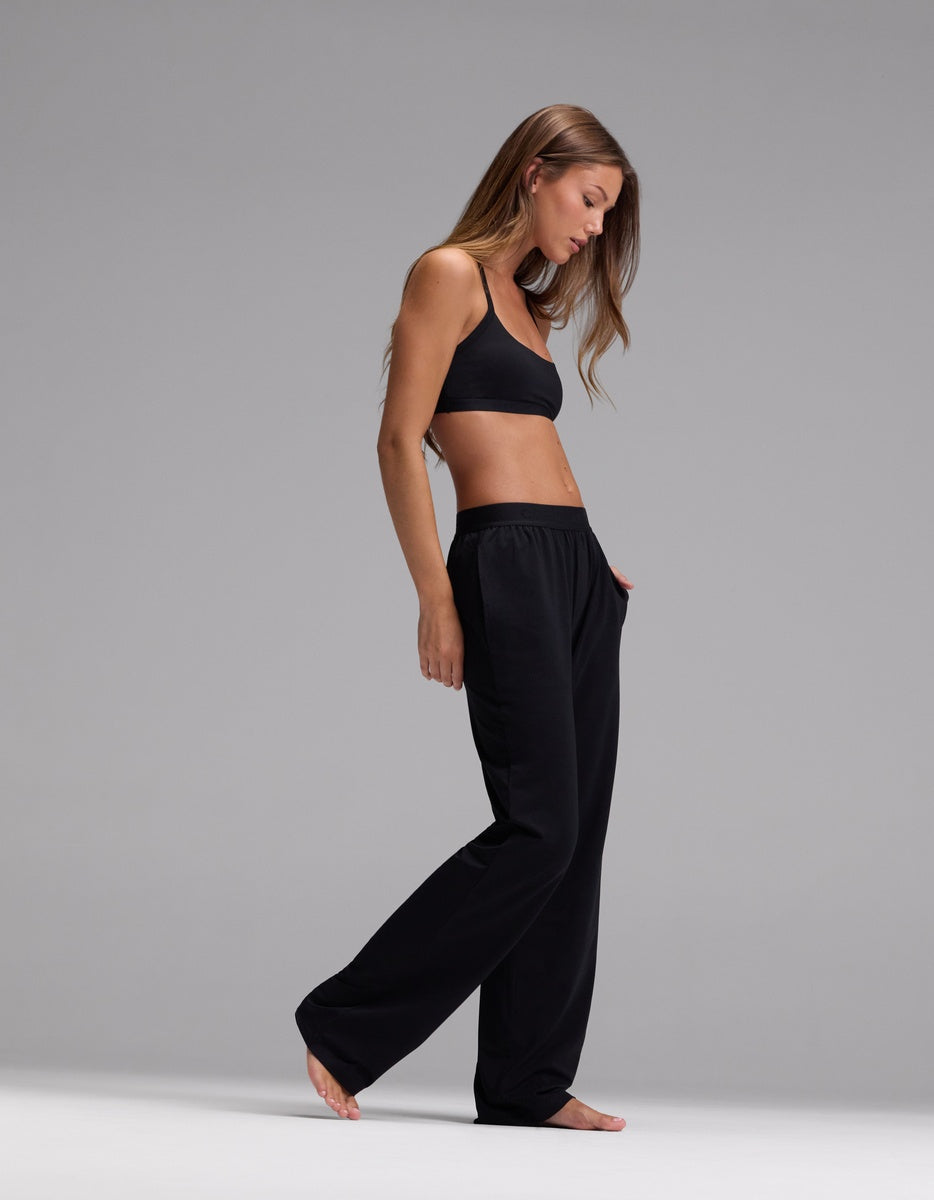 A woman wearing the Cozy Earth Women’s First Layer Pajama Soft Bra & Pajama Pant Set in black stands barefoot against a plain gray background, her long hair down as she looks downward with one hand in her pocket.