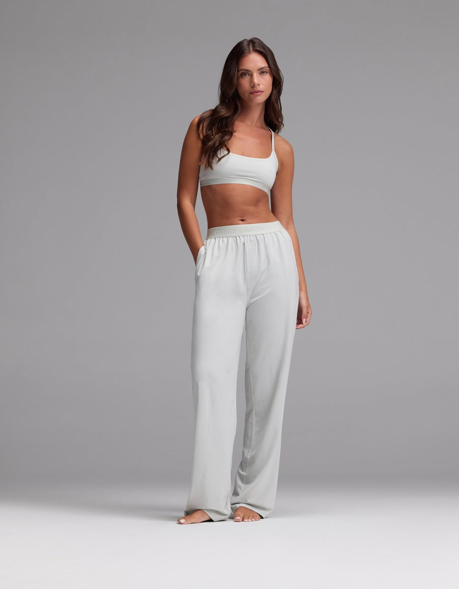 A woman with long brown hair stands barefoot against a plain gray background, wearing the Cozy Earth Women’s First Layer Pajama Soft Bra & Pajama Pant Set in Laurel.