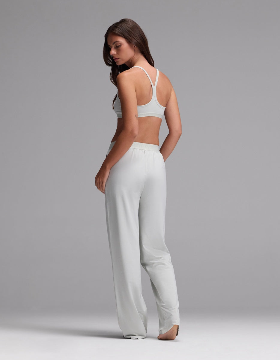 A woman stands barefoot on a gray background, facing away. She wears the Cozy Earth Women’s First Layer Pajama Soft Bra & Pajama Pant Set in a light hue, gazing over her shoulder with her left hand resting at her side.