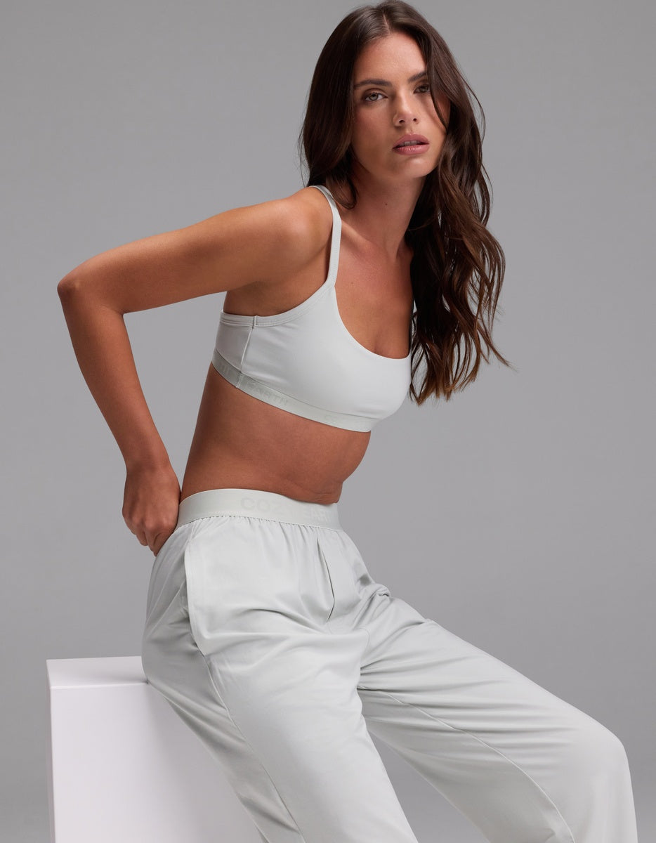 A woman with long brown hair sits on a white block against a plain gray background, wearing the Cozy Earth Women’s First Layer Pajama Soft Bra & Pajama Pant Set in light gray. She gazes slightly upward with a relaxed expression.