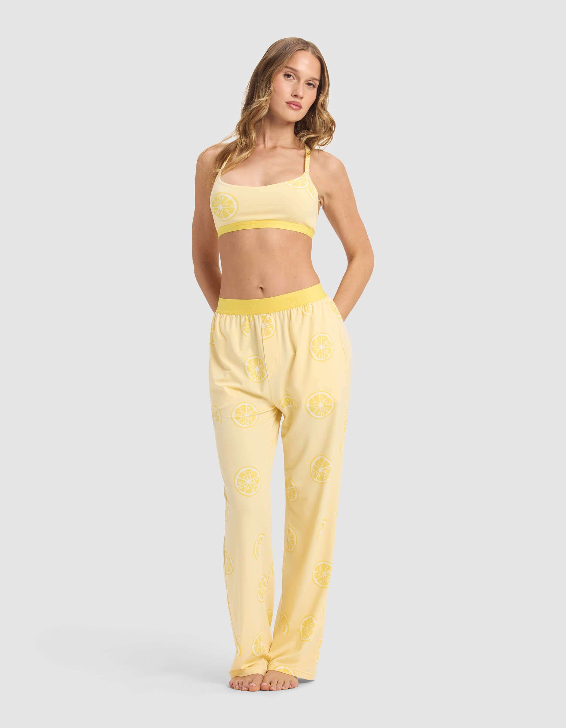 A woman with long light brown hair poses barefoot against a plain background, wearing Cozy Earth's Women's First Layer Pajama Soft Bra & Pant Set—a yellow set with a lemon slice pattern, featuring a crop top and loose pants with pockets.|Color:Lemons