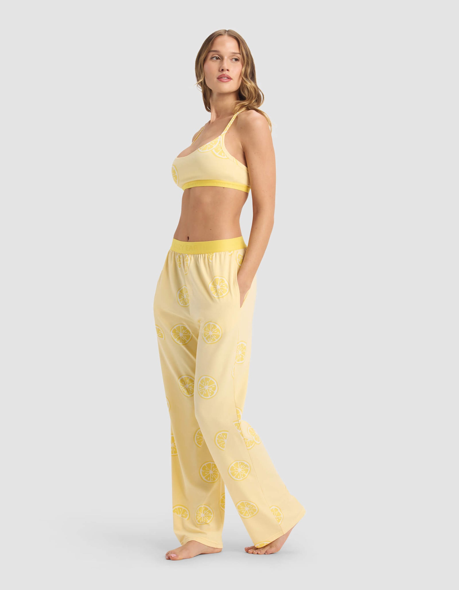 A woman models the Cozy Earth Women's First Layer Pajama Soft Bra & Pant Set in yellow with a lemon slice pattern, standing barefoot against a light gray background with one hand in her pocket and looking over her shoulder.