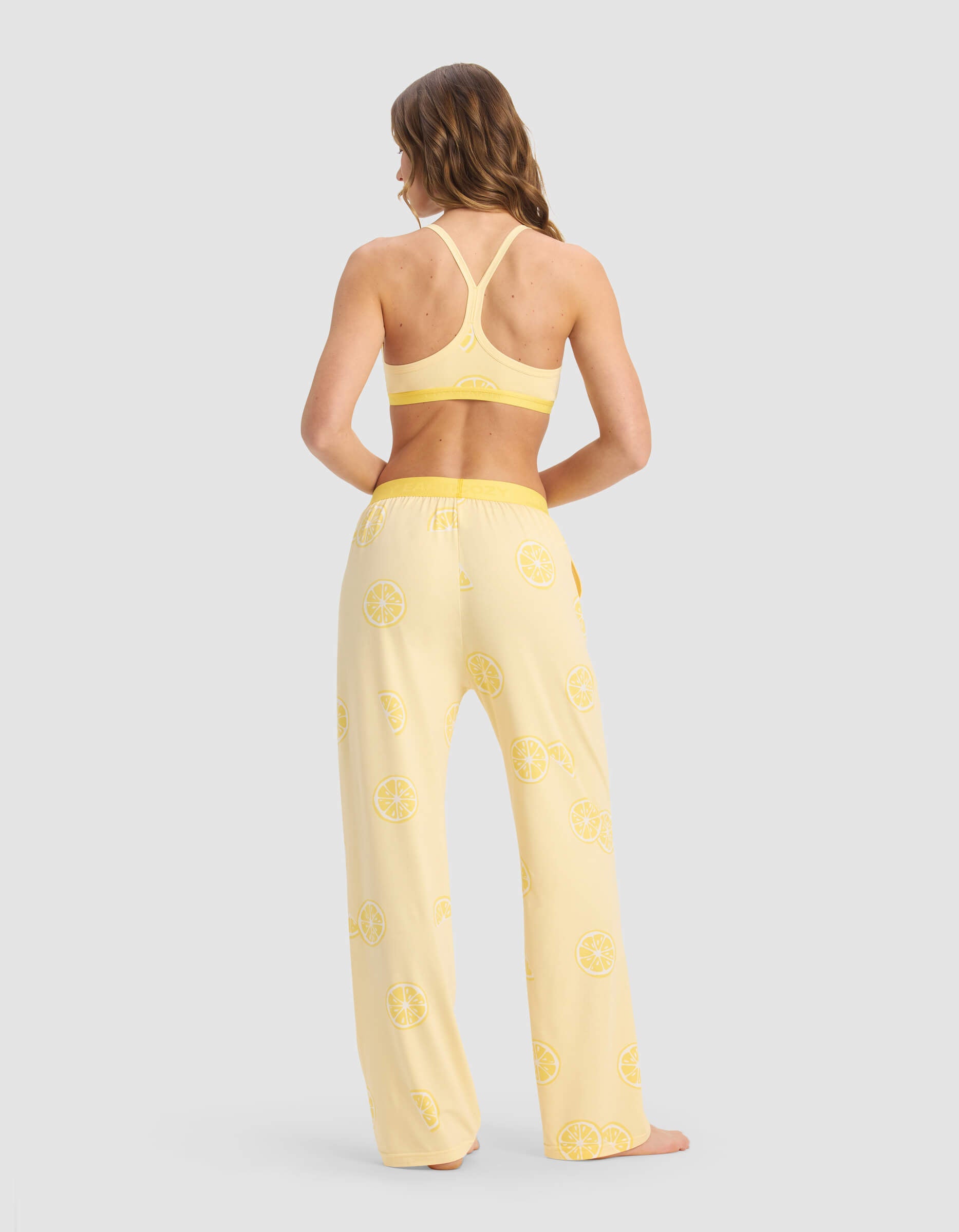 A woman stands barefoot, facing away, wearing the Cozy Earth Women's First Layer Pajama Soft Bra & Pant Set in yellow with lemon slice print on light gray.