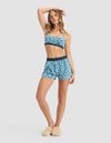 A woman poses in Cozy Earth's Women's First Layer Pajama Soft Bra & Boxer Short Set—blue polka dot loungewear with a dark waistband. She wears beige slippers, one arm on her head, against a plain light gray background.|Color:Blueberries