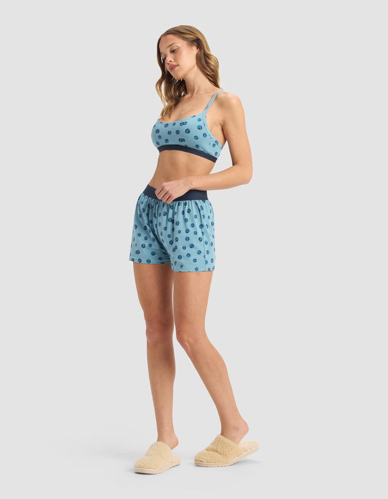 A woman wears the Cozy Earth Women's First Layer Pajama Soft Bra & Boxer Short Set in blue polka dot, paired with cream slippers. Her hair is loose as she looks down, posed against a light grey background.