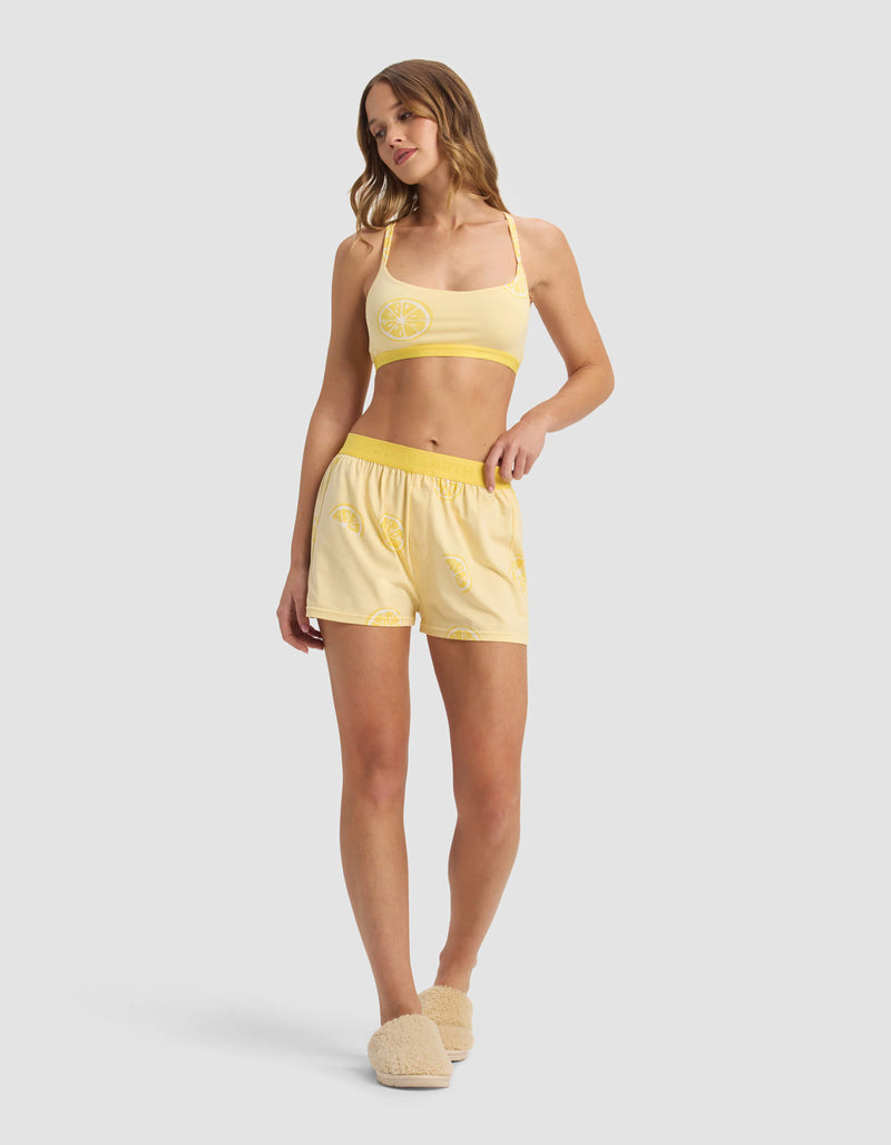 A woman wears the Cozy Earth Women's First Layer Pajama Soft Bra & Boxer Short Set in yellow with lemon prints, paired with beige slippers. She has long wavy hair and poses against a light gray background.