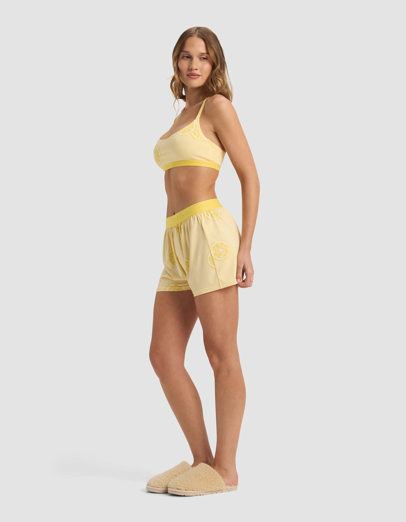 A woman stands sideways in a plain light gray setting, wearing the Cozy Earth Women's First Layer Pajama Soft Bra & Boxer Short Set in yellow with lemon slice designs and beige slippers.