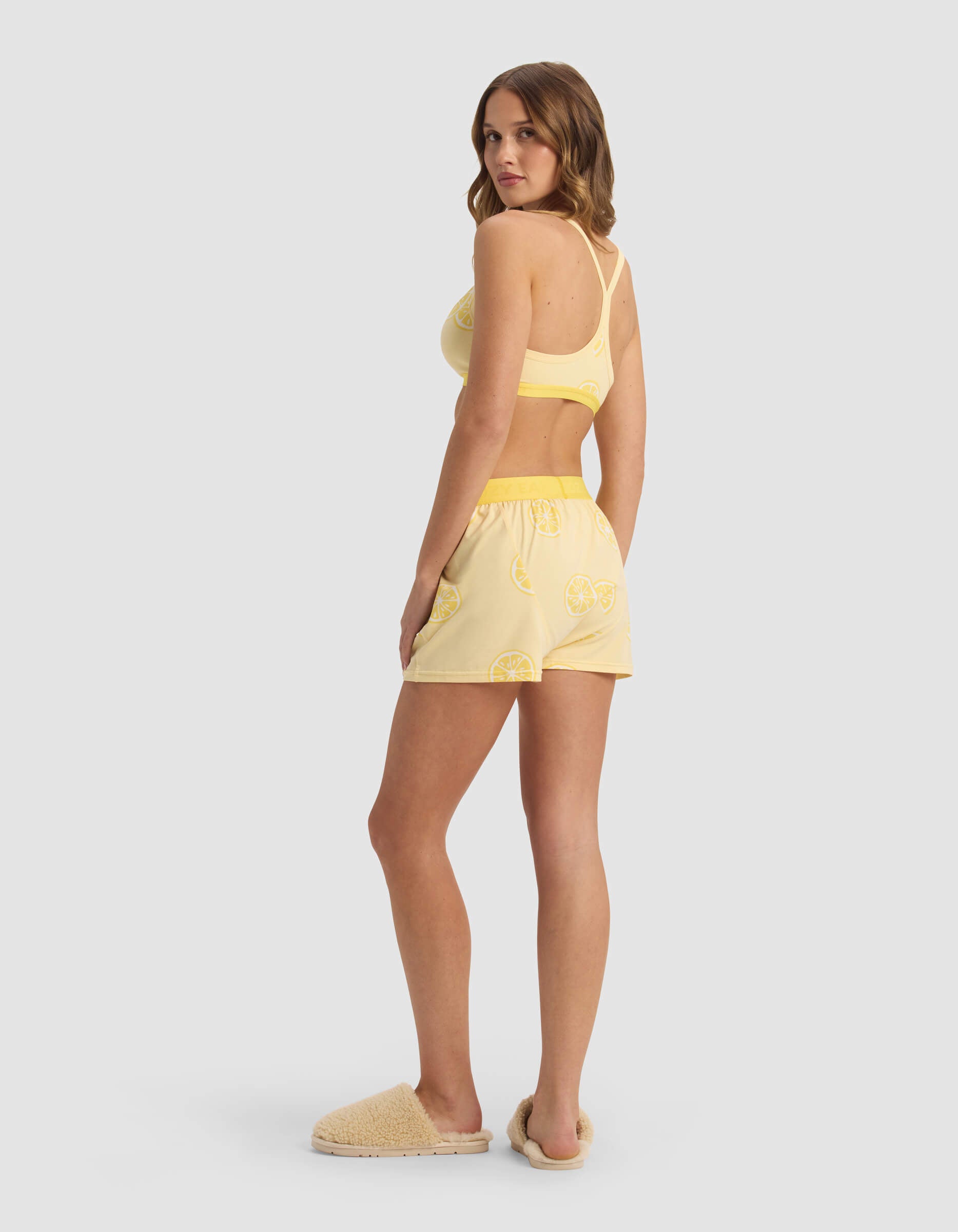 A woman stands sideways, looking over her shoulder, wearing the Cozy Earth Women's First Layer Pajama Soft Bra & Boxer Short Set in yellow with a lemon print and fuzzy slippers, against a plain light gray background.|Color:Lemons