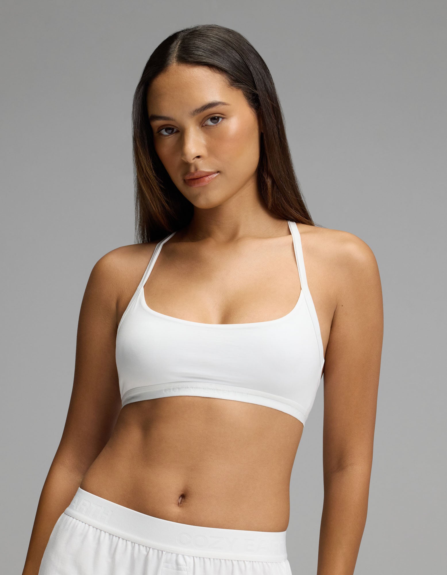 A woman with long brown hair stands confidently against a plain gray background, wearing Cozy Earth’s Women’s First Layer Soft Bra in white paired with white pants.|Color:White