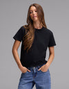 A woman with long brown hair wears the Cozy Earth Women’s Go-To Tee in black and blue jeans, standing with her hands in her pockets against a plain gray background.|Color:Jet Black