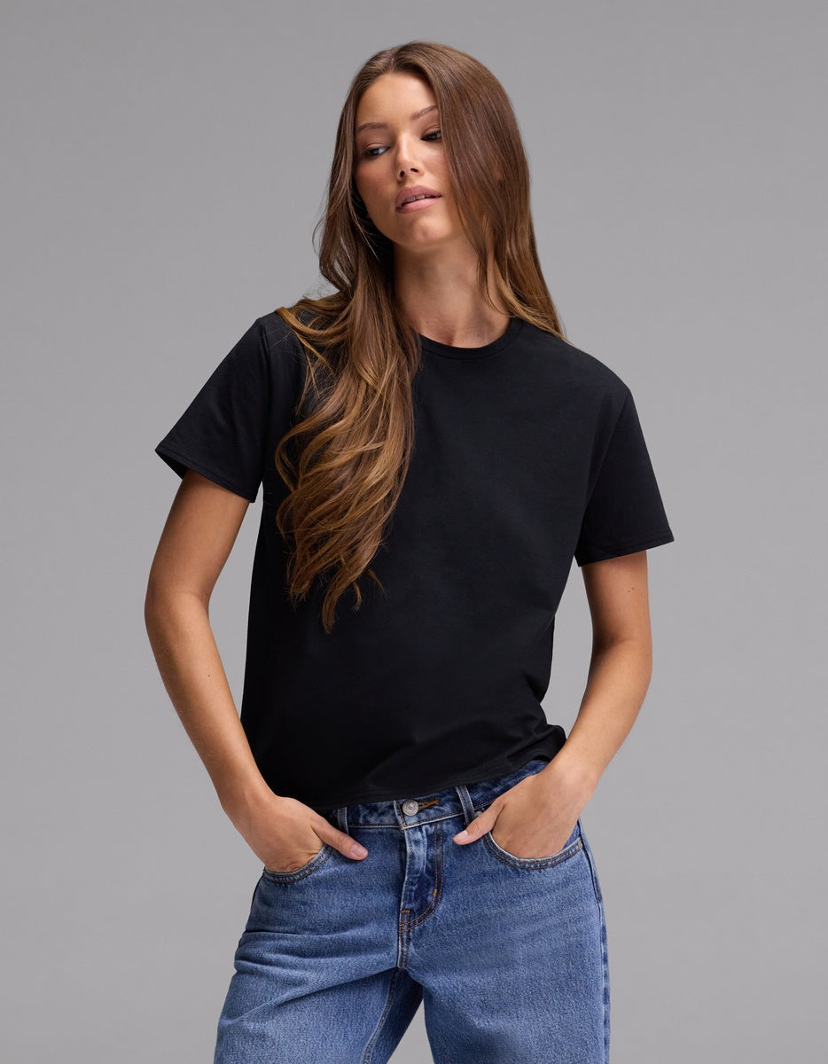 A woman with long brown hair wears the Cozy Earth Women’s Go-To Tee in black and blue jeans, standing with her hands in her pockets against a plain gray background.