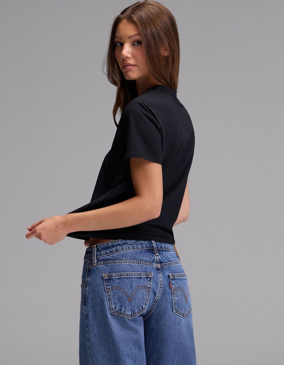 A woman with long brown hair wears the Cozy Earth Women’s Go-To Tee in black and blue jeans, standing sideways and looking at the camera against a plain gray background.