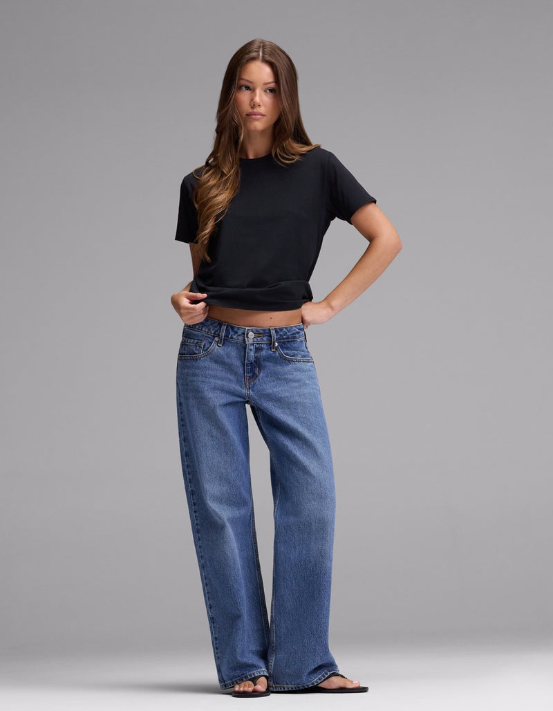 A woman stands against a plain gray background wearing Cozy Earth’s Women’s Go-To Tee in black, paired with blue wide-leg jeans and black sandals. She has long brown hair and a neutral expression, with one hand on her hip.