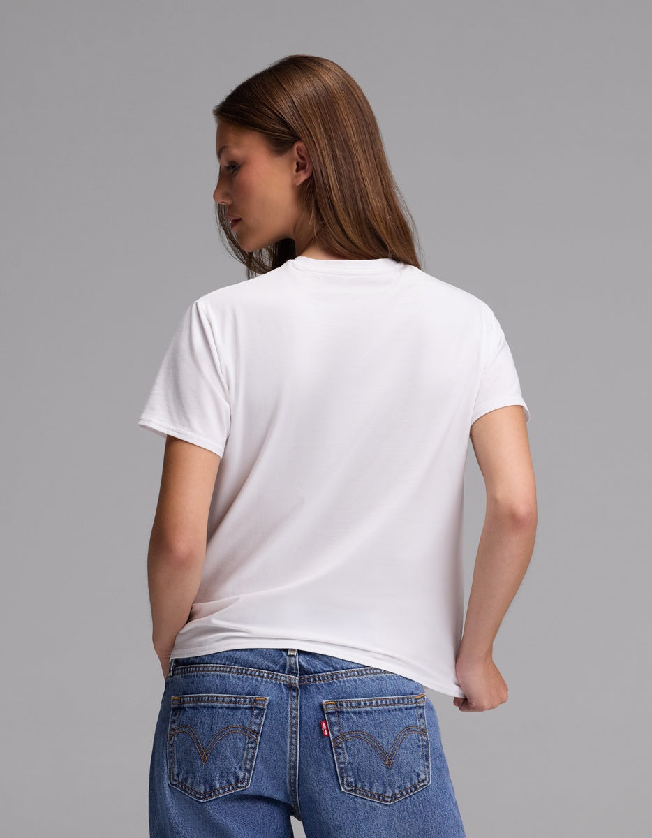 A woman with long brown hair is seen from behind, standing against a gray background while wearing the Cozy Earth Women’s Go-To Tee in white paired with blue jeans.